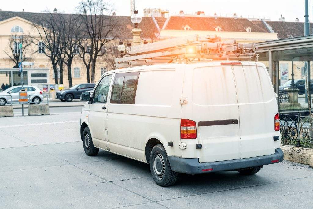 White repair and service van with ladder and orange light bar on roof at city street. Assistance or installation team vehicle.
